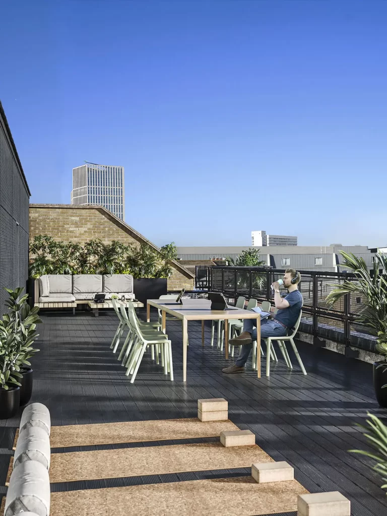 3D render of a rooftop terrace workspace with outdoor seating, a table, and a man working on a laptop.