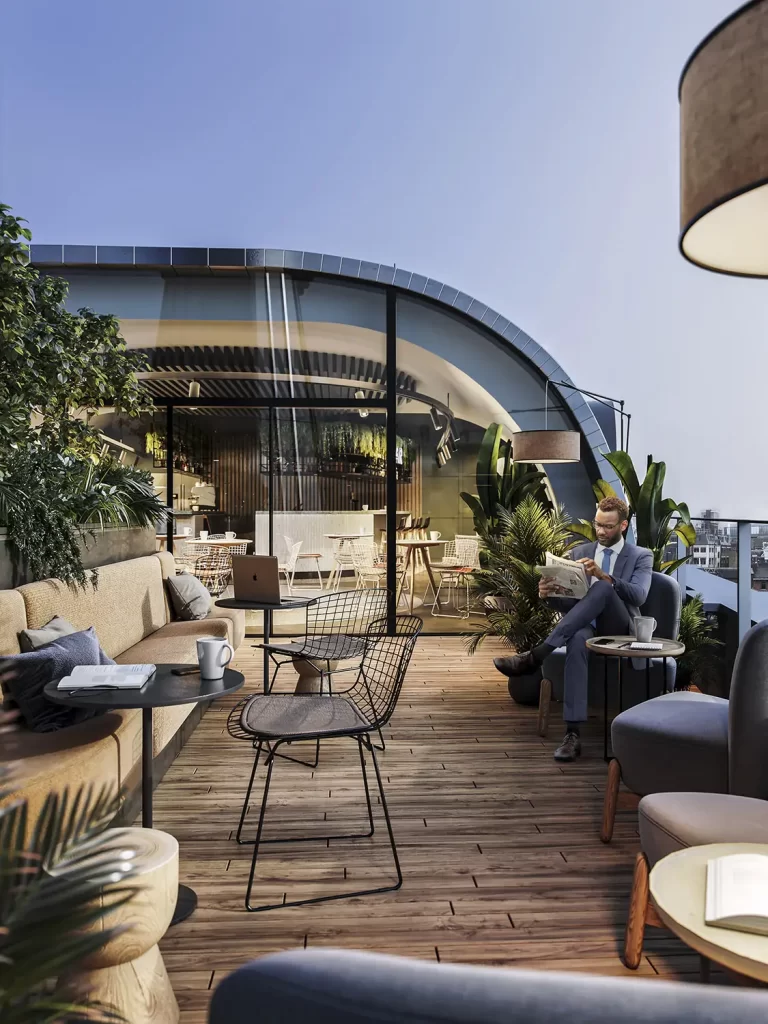 Man in suit relaxing on a rooftop terrace with modern furniture and glass-walled building behind.