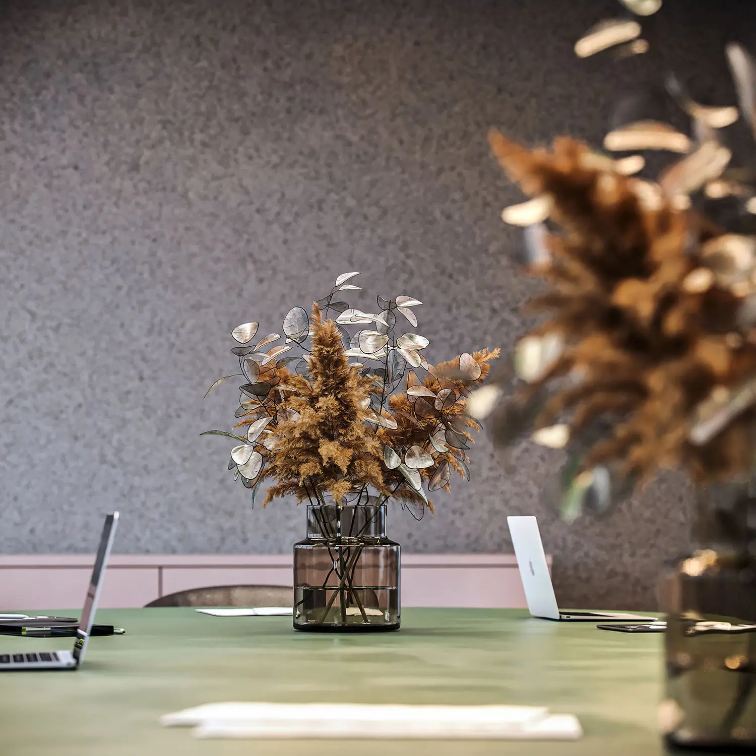 Close-up render of a modern office table with a vase of dried flowers and laptops in the background.